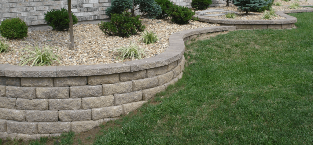 Low tan block retaining wall curving around a gravel planting bed with small shrubs, next to a mown lawn