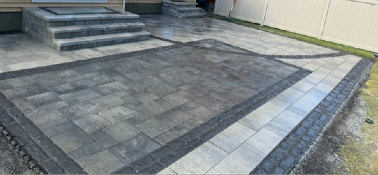 Grey paver patio with a dark patterned border, steps to a rear door and a white privacy fence along one side