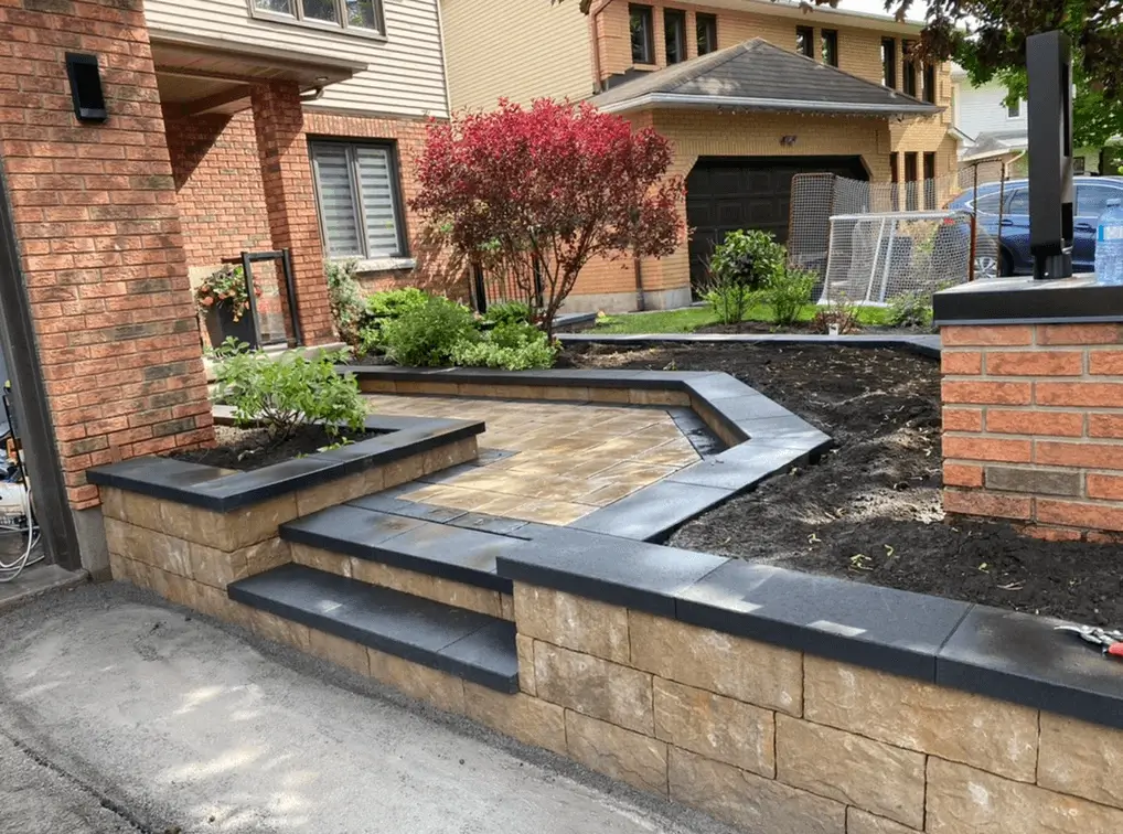 Curved stone block retaining wall with a dark capstone, holding a raised planting bed of shrubs and gravel beside a lawn