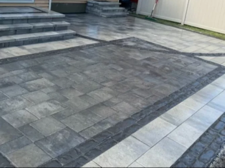 Grey paver patio with a dark patterned border, steps to a rear door and a white privacy fence along one side