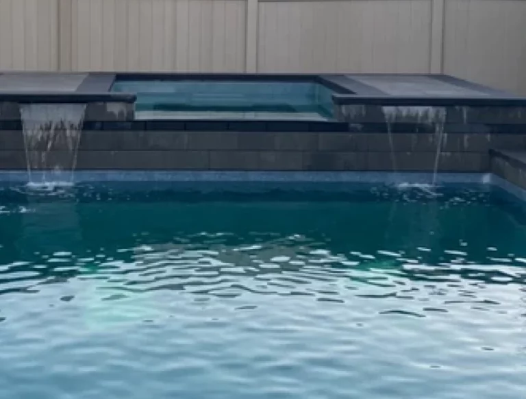 Backyard pool with a raised spa spilling water over its edge, in front of a light privacy fence