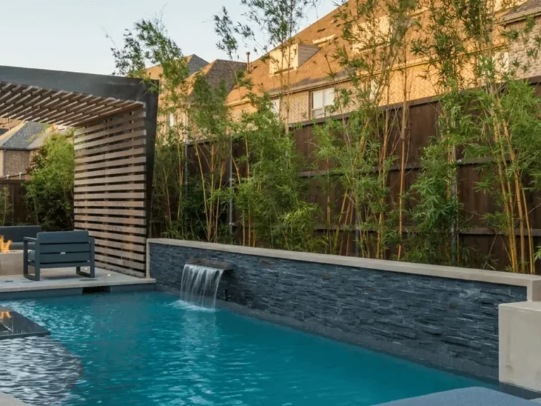 Poolside terrace at dusk with a fire feature, slatted wood pergola, stone-faced water wall and tall screening bamboo